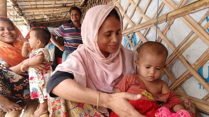 A woman in a pink headscarf cradles her child and plays with a pink toy in a bamboo hut, while others look on