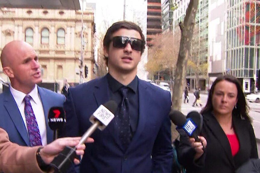 Benjamin wears dark sunglasses, a navy suit and black shirt and is surrounded by reporters.