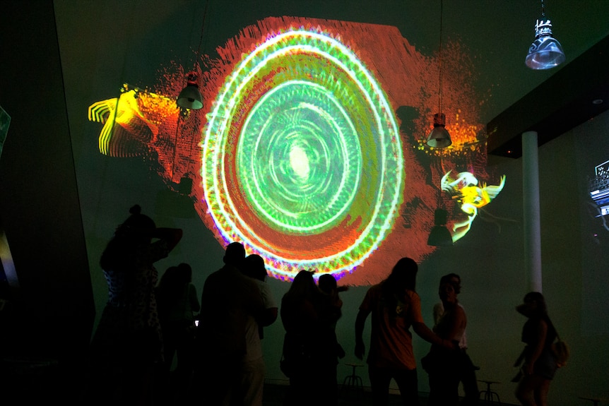 A bright green, red, pink and orange olive-shaped project on a wall, people's silhouettes in the foreground.