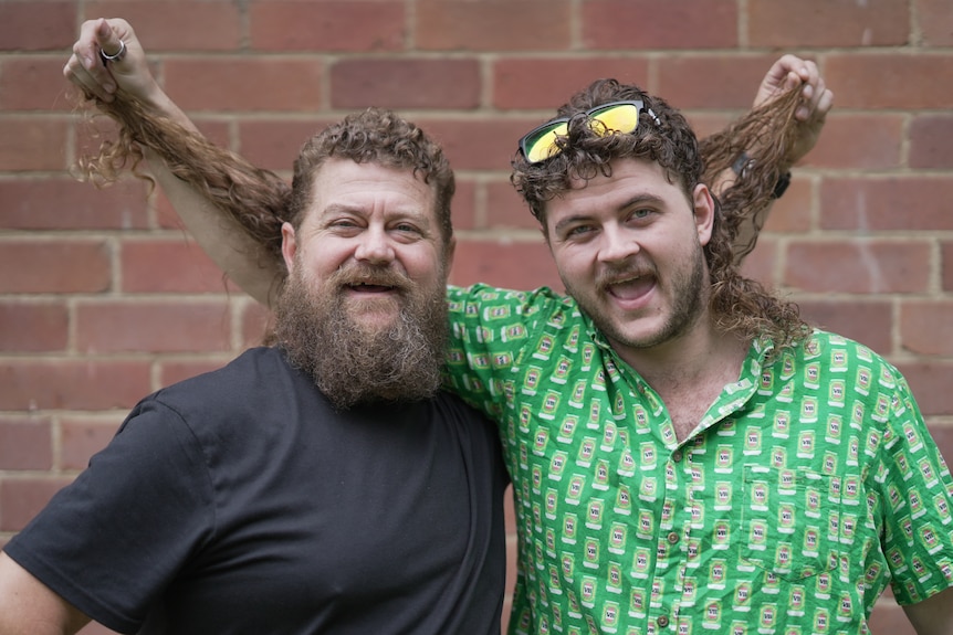 Two men standing side by side lifting each other's mullets.