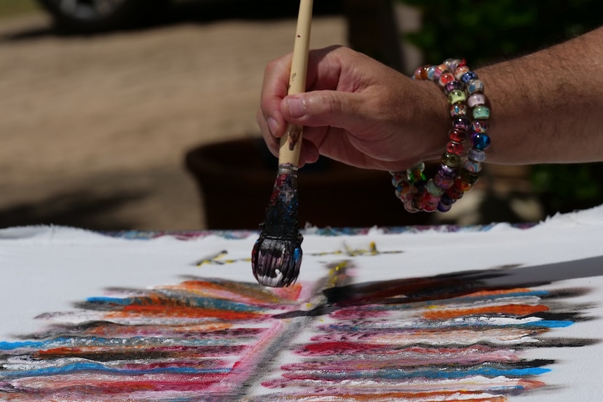 A man with beaded bracelet around his wrist holding a black paintbrush over a white dress he is painting