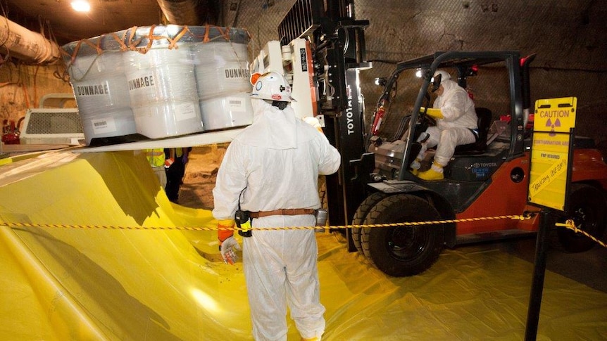 A man in a hazmat suit looks on as another man operates a forklift with vats of toxic waste