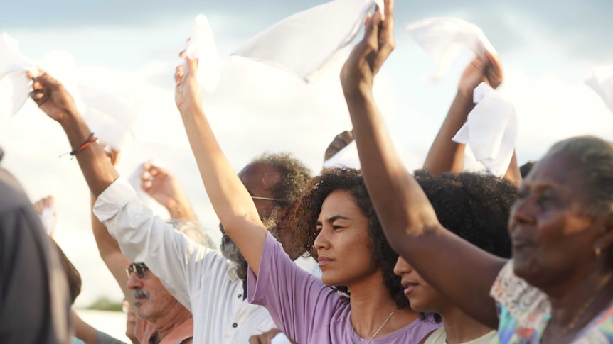 Side on view of line of six people with their right arms held in air, and their right hands holding white handkerchiefs.