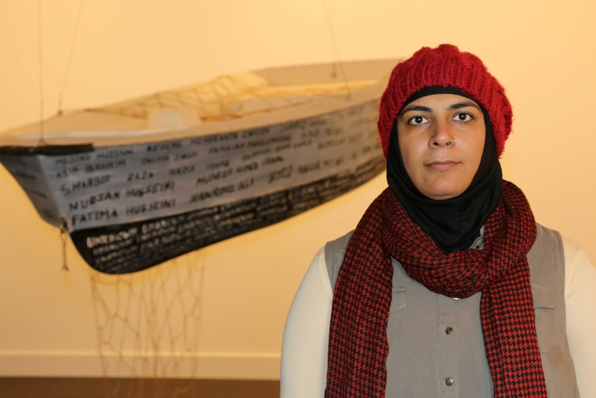 Artist Marziya Mohammedali stands in front of her work, a wooden dinghy hanging from the ceiling with words on it.