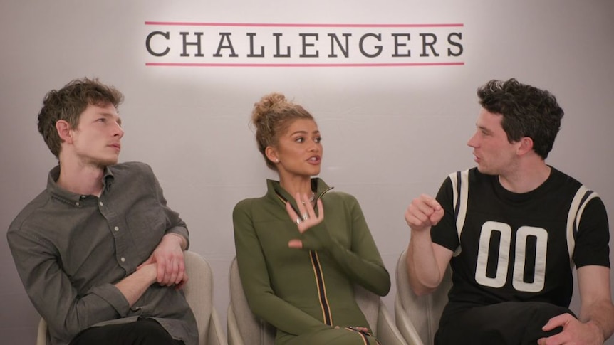 A woman speaks to a man during a press junket. Another man listens next to them. 'Challengers' is written above them.