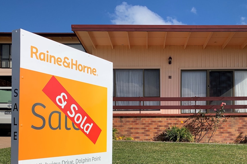 A sold sticker across a Raine & Horne sign on a lawn outside a small house.