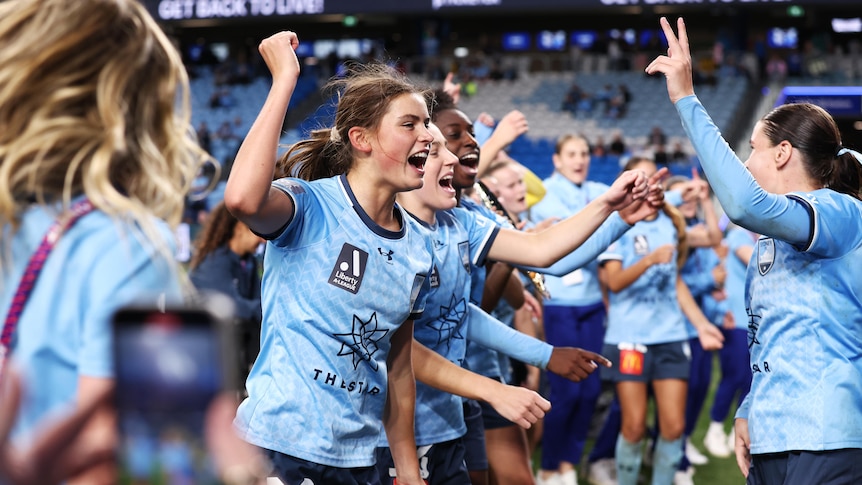 A women's soccer team wearing light blue jerseys celebrate with their arms in the air after a game