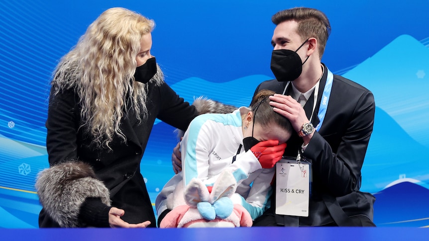 A female figure skater covers her face with her hand as she cries while her coach and choreographer console her.