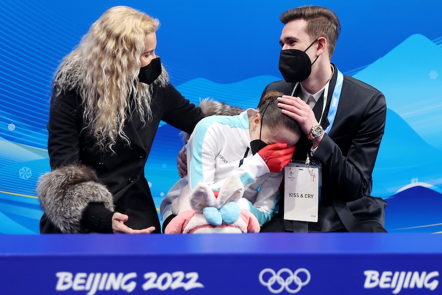 A female figure skater covers her face with her hand as she cries while her coach and choreographer console her.