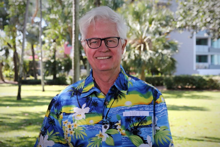Un homme avec des lunettes dans une chemise hawaïenne colorée sourit dans un parc rempli de palmiers