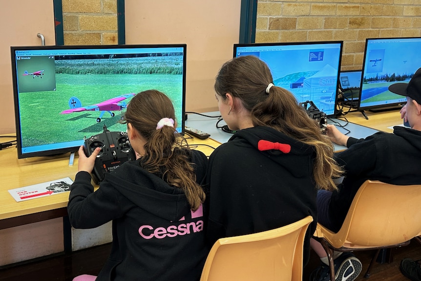 Two girls facing computers, back to camera, hold remote controls and playing on a flight simulator.