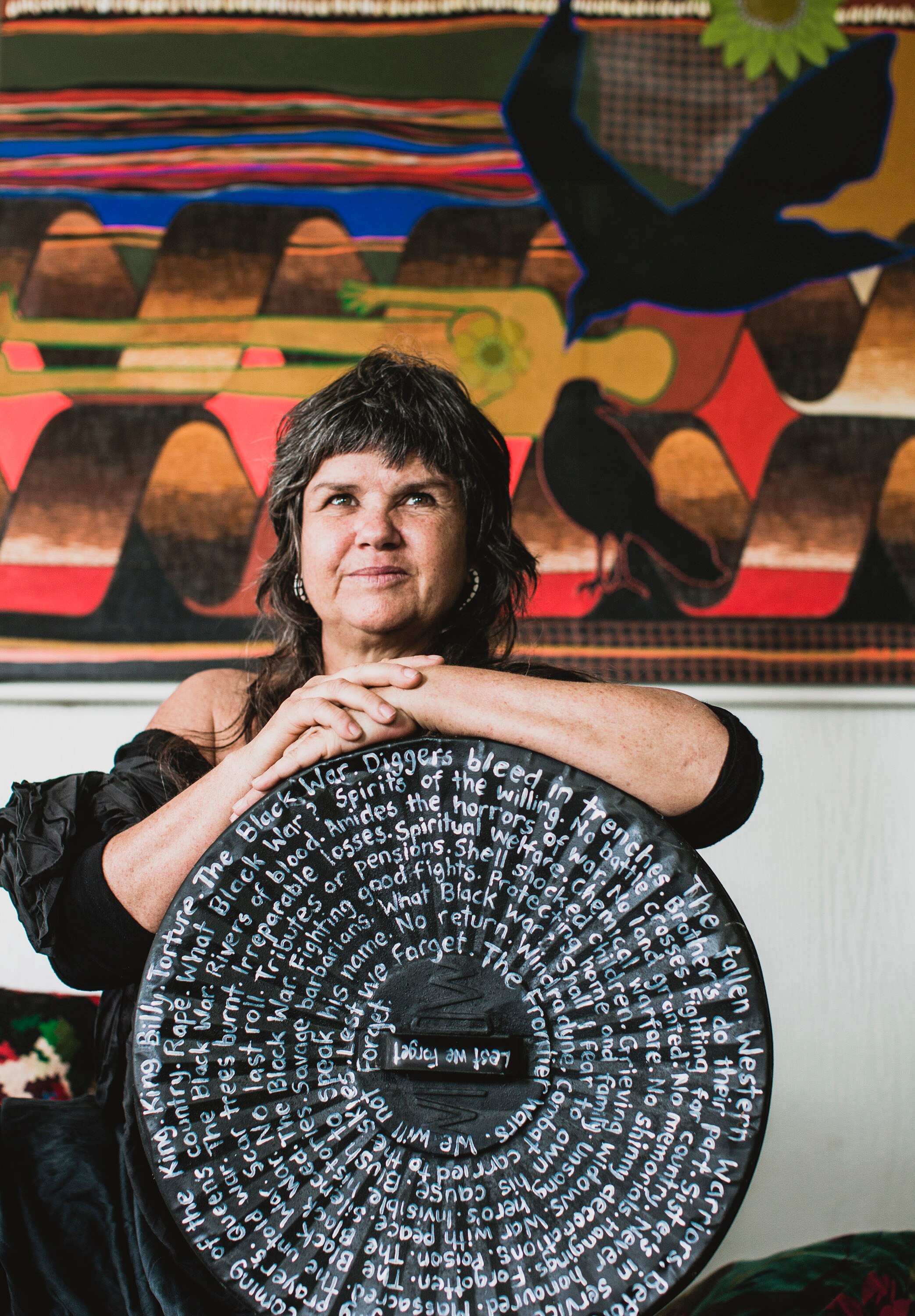 A 50-something Aboriginal woman with a mullet sits with her arms resting on a circular sculpture, handpainted with words