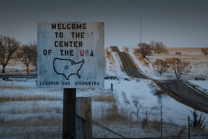 A sign reads welcome to the centre of the USA, beside a snowy road.