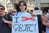 A sign at international women's day march reads "I object", with "am a" crossed out.