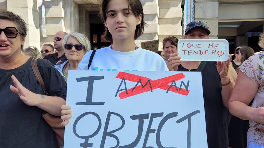 A sign at international women's day march reads "I object", with "am a" crossed out.
