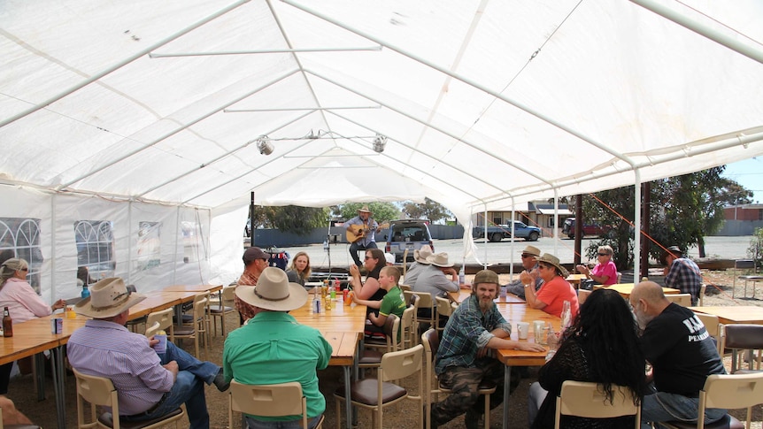 Dozens of people sit under a white marquee at tables and watch a country music singer perform