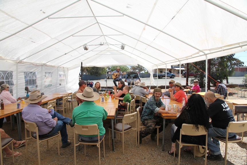 Dozens of people sit under a white marquee at tables and watch a country music singer perform