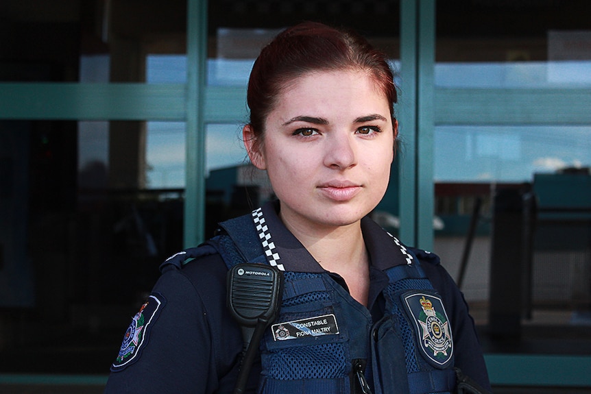 A woman in police uniform looks straight at the camera, standing beside a police 4WD vehicle.