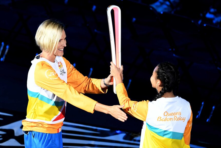 A woman with short blonde hair wearing a gold tracksuit top hands a tall baton to a young girl.