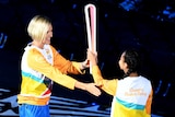 A woman with short blonde hair wearing a gold tracksuit top hands a tall baton to a young girl.