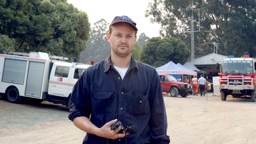 Vincent in fire gear holding microphone and recorder standing in front of fire vehicles in street.