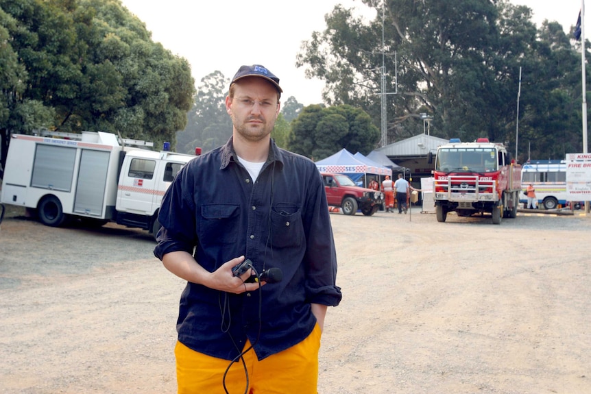Vincent in fire gear holding microphone and recorder standing in front of fire vehicles in street.