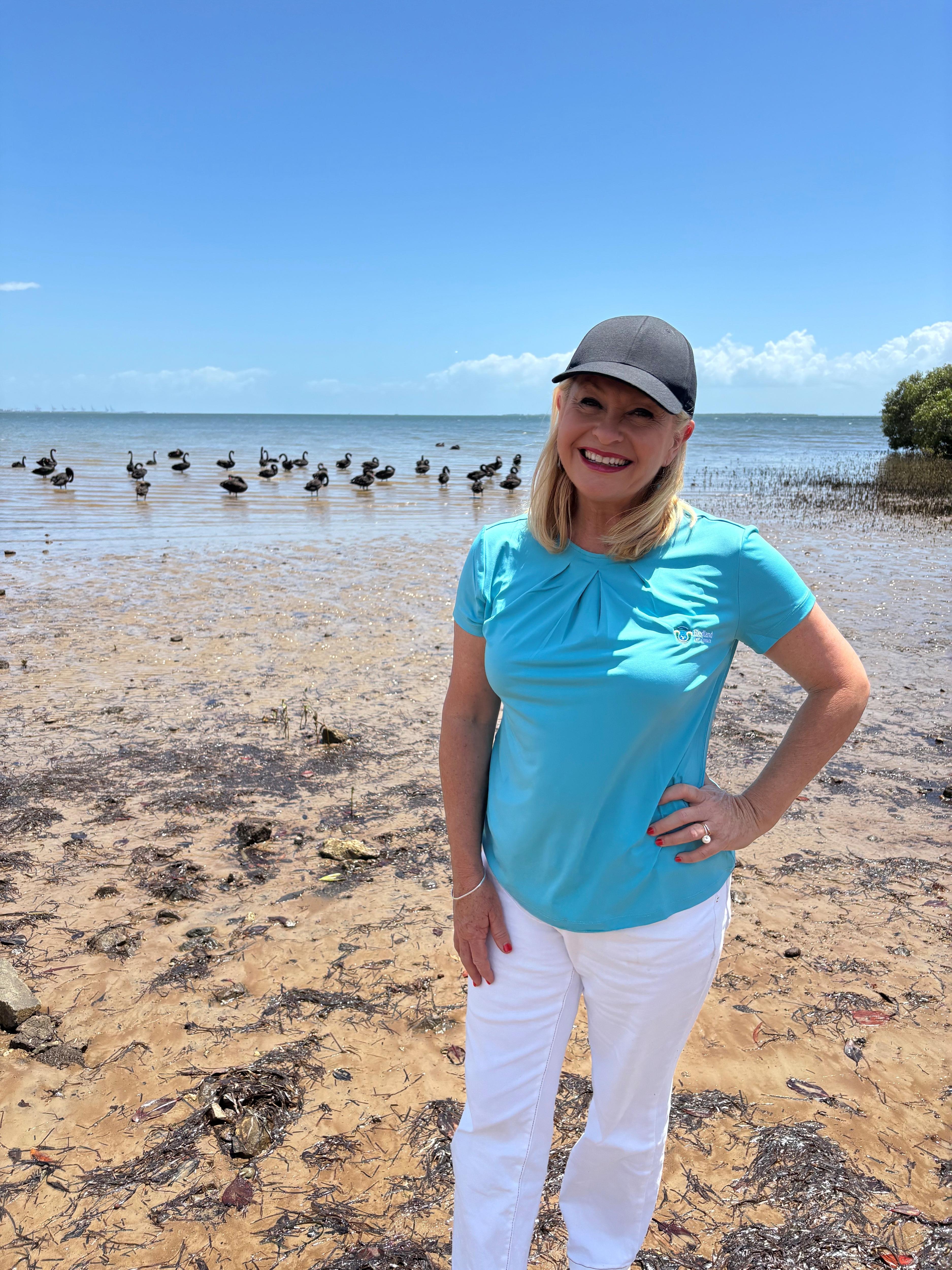 A blonde woman in a blue t-shirt standing on the beach with a flock of black swans behind her, she is smiling at the camera.