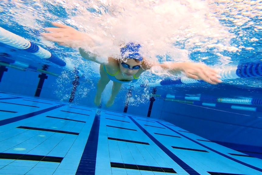 An underwater photo of a teenage girl with a swim cap and goggles swimming butterfly in a pool.