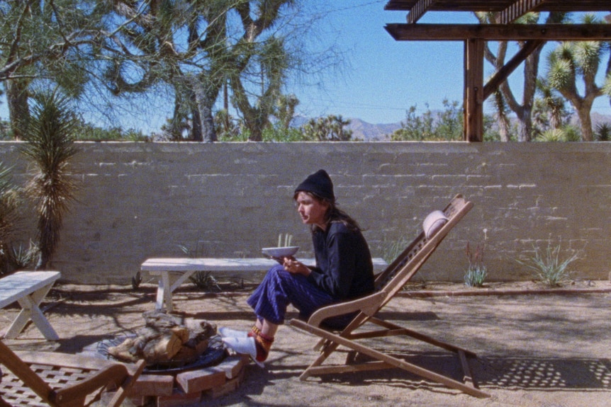 A white woman with shaggy brown hair is wearing a black shirt, blue pants and a black beanie and is sitting on a deck chair in the yard.