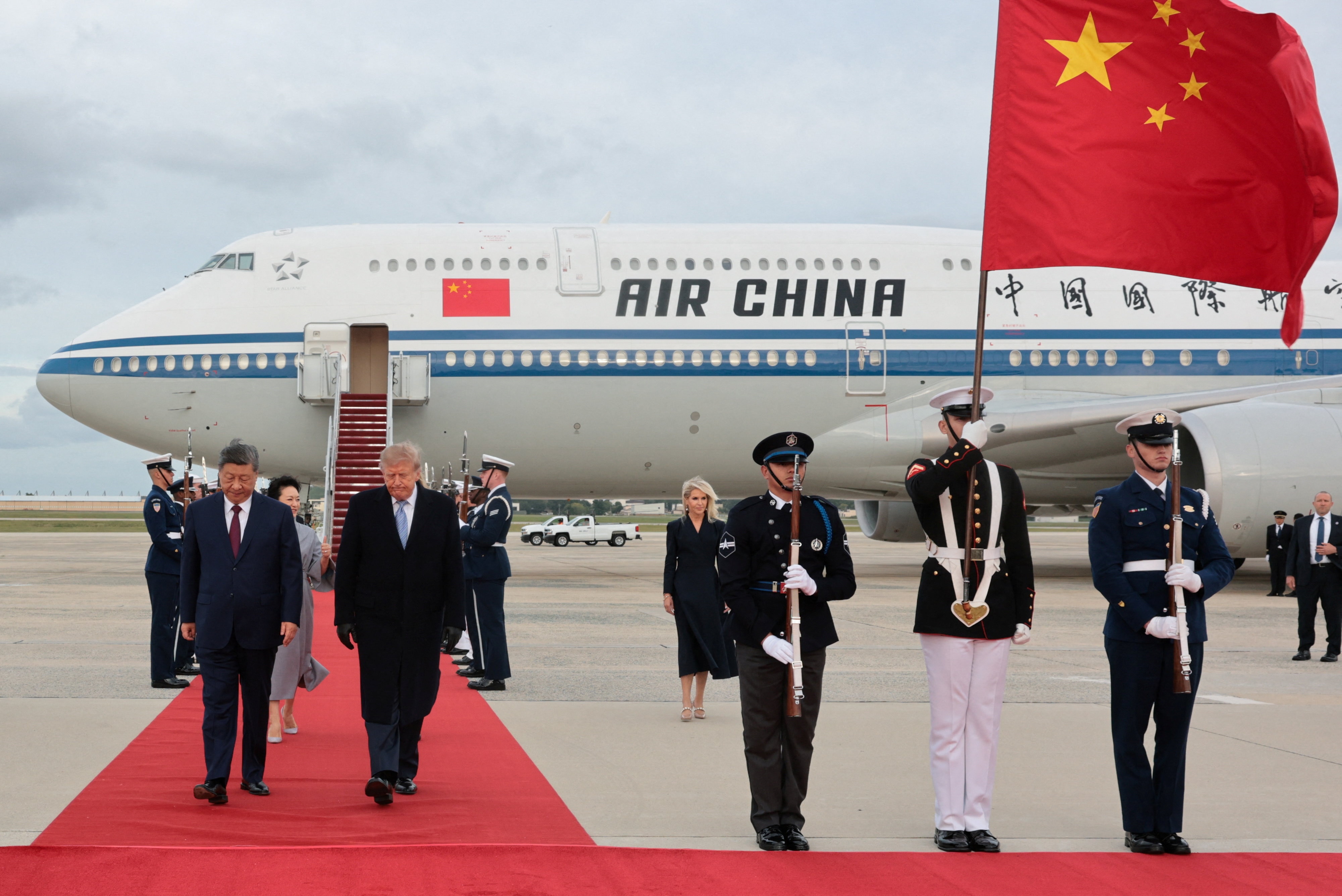 Donald Trump and Xi Jinping walk down a long, red carpet laid out on the tarmac, with a large jumbo jet in the background.