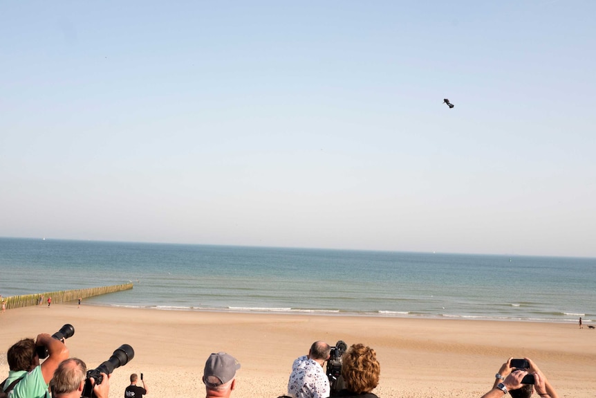 A group of people stand on a beach taking pictures as a man on a hoverboard is seen in the blue sky. The sea is below.