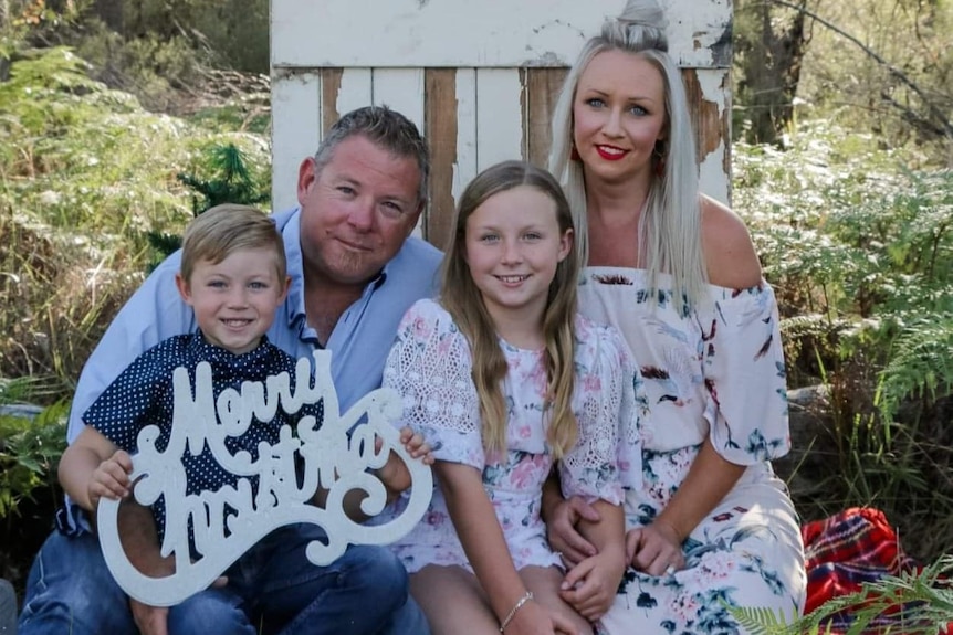 Ballandean volunteer firefighter Aaron Cox with his wife Bindi and two children sitting on rug with Merry Christmas sign.