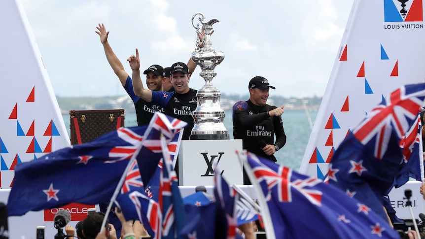 Four Team NZ sailors stand on a podium with a multi-tiered silver trophy in front of NZ-flag-waving crowd