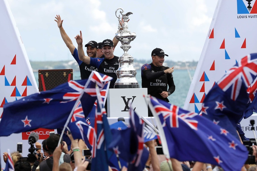 Four Team NZ sailors stand on a podium with a multi-tiered silver trophy in front of NZ-flag-waving crowd