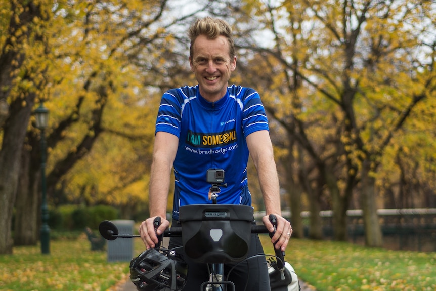 man sitting on a bike in a park with a big smile on his face, wearing a blue top