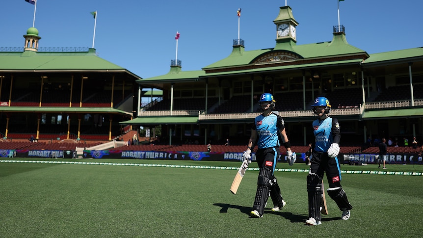 Two Adelaide WBBL cricketers walk on to the SCG, with an old pavilion behind them and blue sky overhead. 