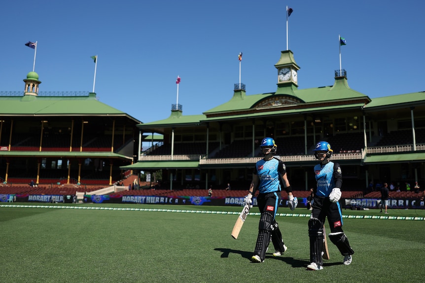 Two Adelaide WBBL cricketers walk on to the SCG, with an old pavilion behind them and blue sky overhead. 