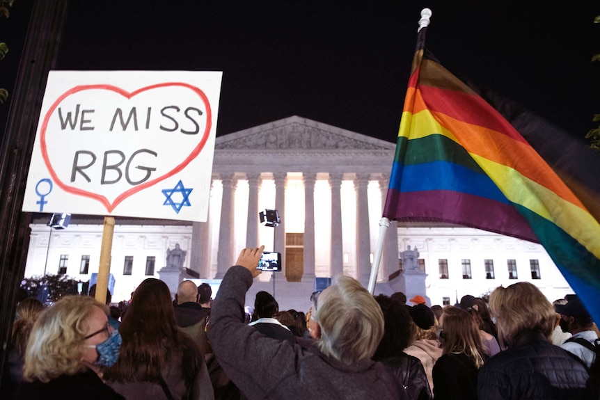 People gather out the front of the US Supreme Court. One person is holding a sign that says 'we miss RBG' and a rainbow flag