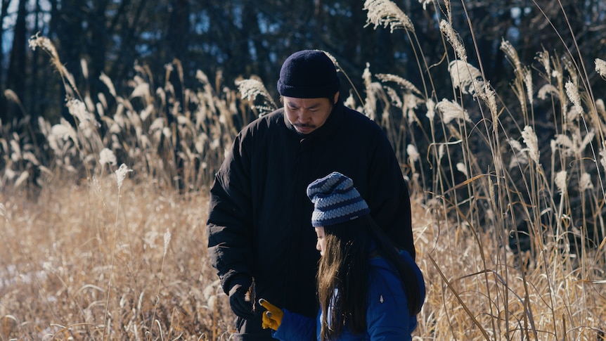 A man in black and a girl in a beanie and blue coat stand in a wheat field, the wheat standing tall above the man.