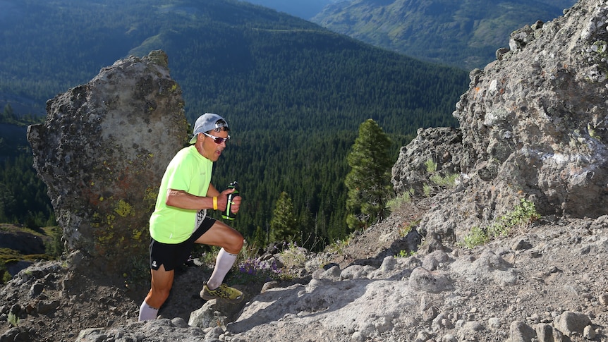 Man dressed in black shorts, yellow t-shirt and backwards cap runs up a rocky slope holding a drink bottle.