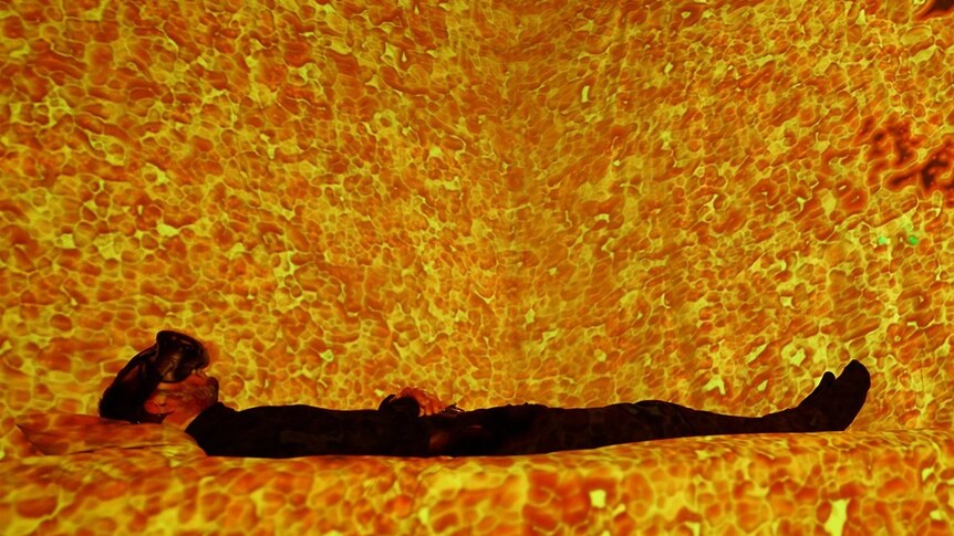 Participant lies in interactive bed wearing VR headset, surrounded by kaleidoscopic visuals.