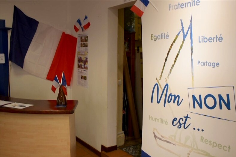 In a dimly lit reception area, you view lots of French flags with a large sign that that reads 'mon non est'.