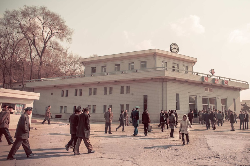 A desaturated image shows a crowd of North Koreans in dark clothing approach a teal subway station.