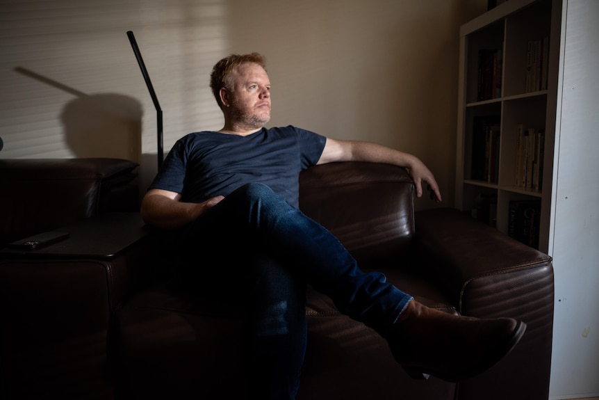 A man sits on a couch, looking to the side with a serious expression.