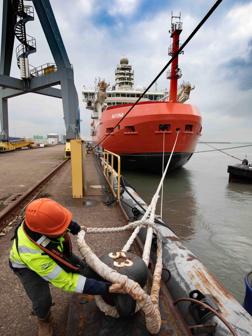 A worker removes a rope on a dock with an orange ship in the background.