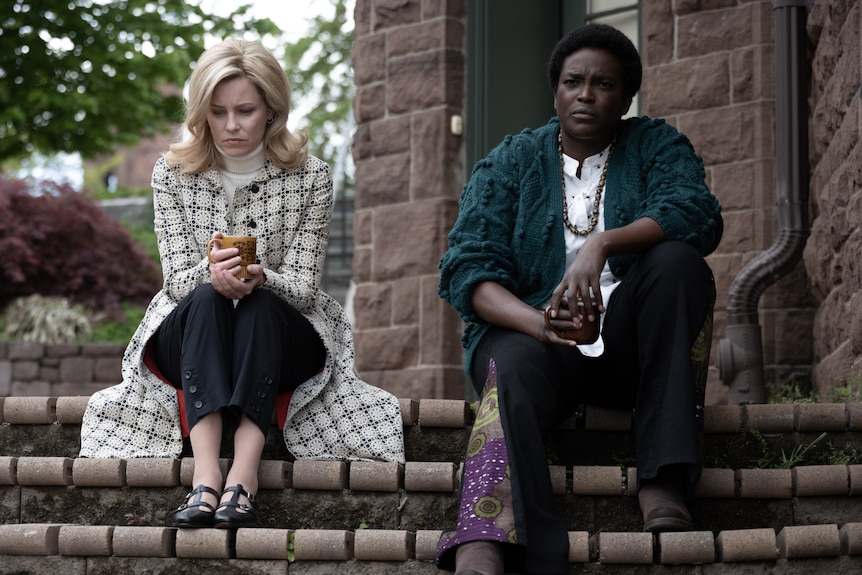 A blonde middle-aged woman and a Black woman sit on a set of outdoor steps. They both look concerned and upset.