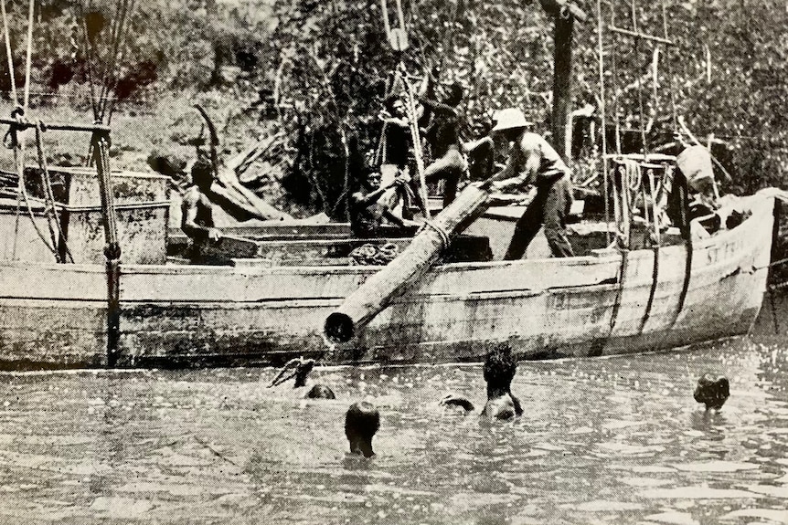 A black and white image of a wooden boat carrying a tree log. Aboriginal men are in the water helping to move the timber 