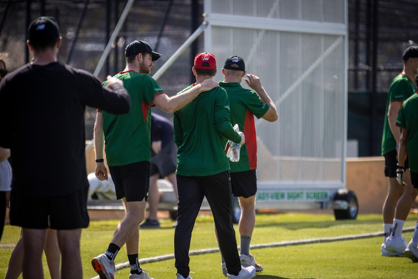 A group of men walk out on to a cricket ground, one with his hand on the shoulder of another