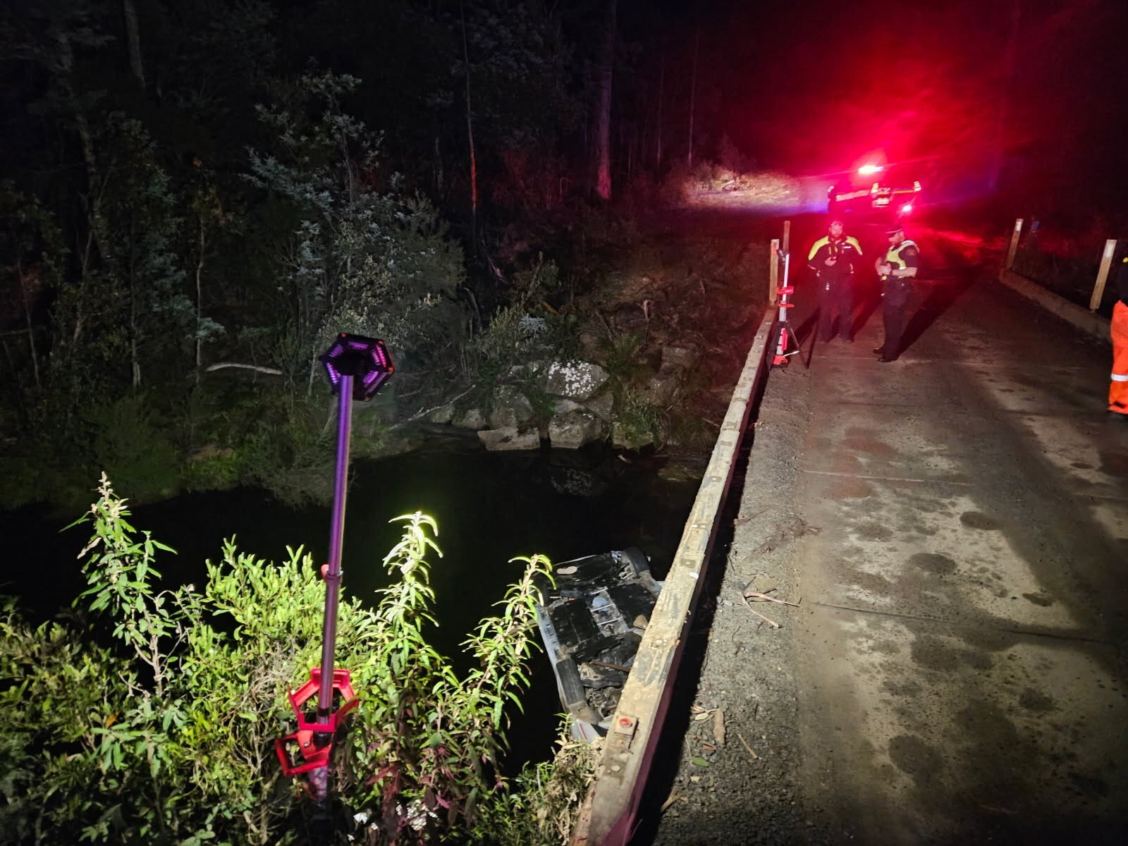Police stand on a concrete bridge where a car is upside down in the river below