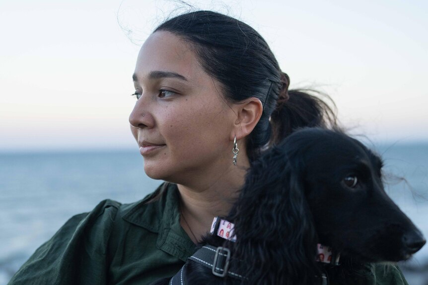 A woman holds a black dog overlooking the beach.